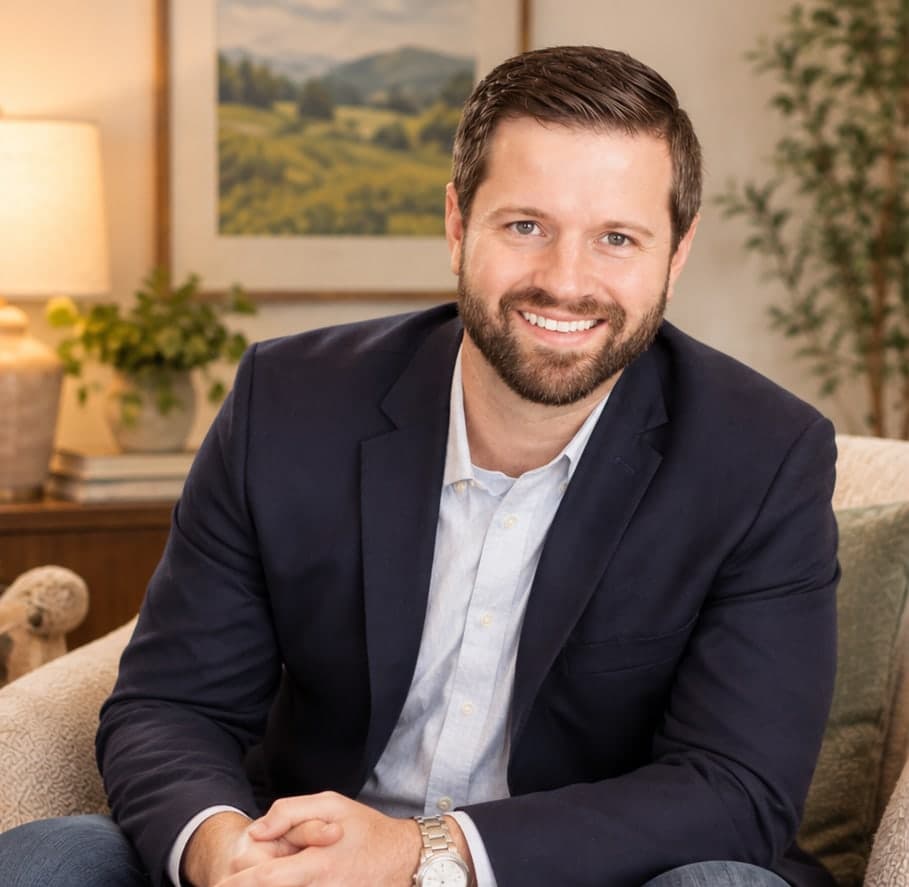 Jason Newton, smiling, with a beard, in a navy blazer and light blue shirt, seated in an armchair